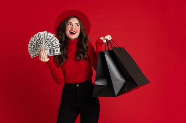 Beyaz kadın kırmızı arka planda izole edilmiş alışveriş torbaları ve dolarlarla poz verirken gülüyor.