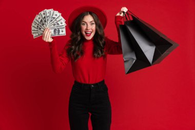 Beyaz kadın kırmızı arka planda izole edilmiş alışveriş torbaları ve dolarlarla poz verirken gülüyor.