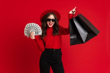 Beyaz kadın kırmızı arka planda izole edilmiş alışveriş torbaları ve dolarlarla poz verirken gülüyor.