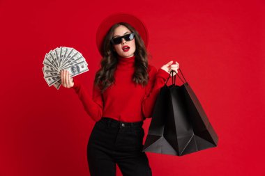 Güneş gözlüklü beyaz kadın, kırmızı arka planda izole edilmiş alışveriş torbaları ve dolarlarla poz veriyor.