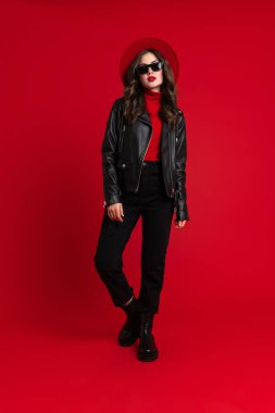 Deri ceketli beyaz kadın kırmızı arka planda yalnız başına poz veriyor ve kameraya bakıyor.