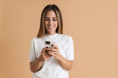 Tişört giyen, gülümseyen ve bej arka planda izole cep telefonu kullanan beyaz bir kadın.