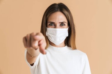 Yüzü maskeli beyaz kadın bej arka planda izole edilmiş kamerayı işaret ediyor.