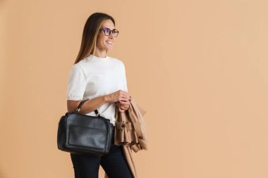 Gözlüklü beyaz kadın, bej arka planda izole edilmiş çantayla poz verirken gülümsüyor.