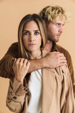 Beyaz sarışın çift, bej arka planda yalnız poz verirken birbirlerine sarılıyorlar.