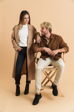 Bej arka planda birlikte poz verirken gülümseyen beyaz sarışın çift.