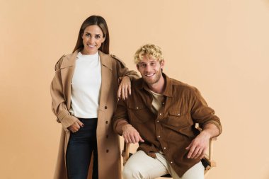 Beyaz sarışın çift kameraya gülümserken bej arka planda yalnız poz veriyorlar.