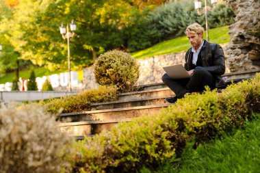 Sarışın adam dışarıda taş bir bankta otururken dizüstü bilgisayarla çalışıyor.