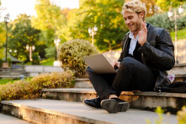 Sarışın adam dışarıda taş bir bankta otururken dizüstü bilgisayarla çalışıyor.