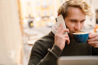 Sarışın kıllı adam açık kafede otururken kitap okuyor.
