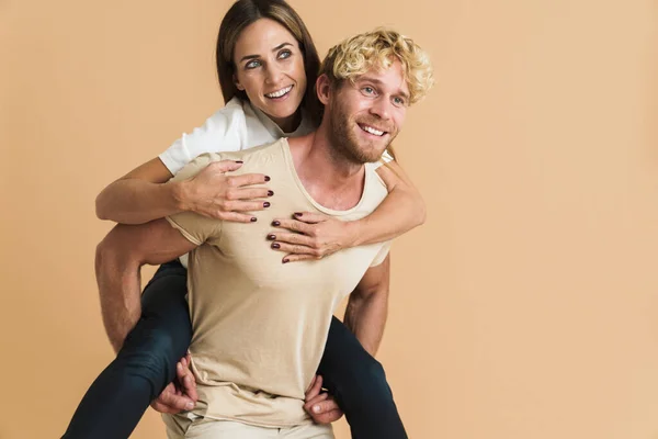 White couple wearing t-shirts smiling while piggybacking isolated over beige background
