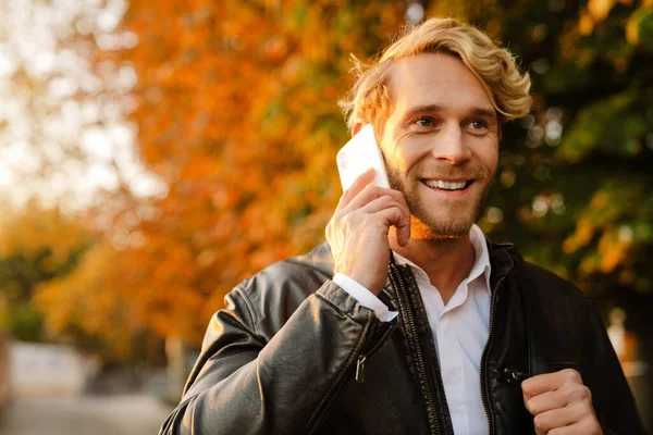 Blonde man smiling and talking on mobile phone while walking in autumn park