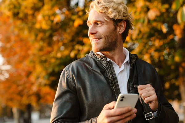 Blonde man smiling and using mobile phone while walking in autumn park