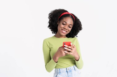 Saçbandı takan, gülümseyen ve cep telefonu kullanan genç siyah bir kadın beyaz arka planda izole edilmiş.