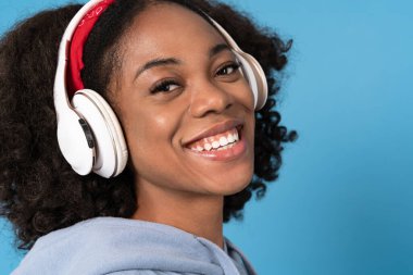 Mavi arka planda izole edilmiş kulaklıklarla müzik dinlerken gülümseyen genç siyahi kadın.
