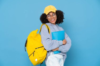 Sırt çantası ve klasörle poz verirken gülümseyen genç siyah kadın mavi arka planda izole edilmiş.