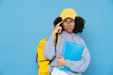Sırt çantası ve klasörle poz veren siyah şaşkın kadın mavi arkaplan üzerinde izole