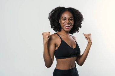 Young black woman in sportswear laughing and holding clenched fists isolated over white background