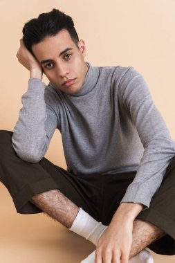 Genç Ortadoğulu adam bej arka planda izole bir şekilde yerde otururken kameraya bakıyor.