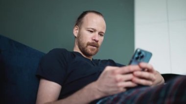 Pijama giyen neşeli sarışın adam evdeki kanepede otururken telefonla yazı yazıyor.
