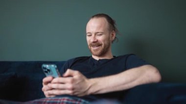 Pijama giymiş gülümseyen sarışın adam evdeki kanepede otururken telefonla yazı yazıyor.