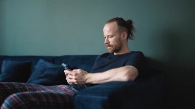 Kendine güvenen, pijama giyen, evde koltukta otururken telefonda yazan sarışın bir adam.