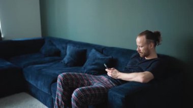 Evdeki kanepede otururken telefonda yazan pijama giyen ciddi sarışın bir adam.