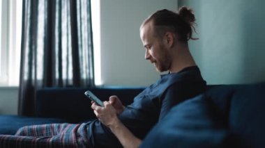 Pijama giyen mutlu sarışın adam evdeki kanepede otururken cep telefonunda mesajlaşıyor.