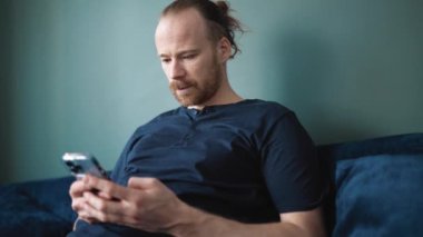 Laughing blond man wearing pajama texting on mobile while sitting on the sofa at home