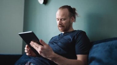 Smiling blond man wearing pajama looking at tablet while sitting on the sofa at home