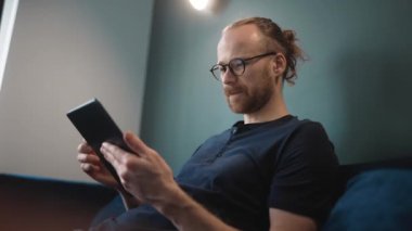 Kendine güvenen, gözlüklü, tablete mesaj atan, evdeki kanepede oturan sarışın bir adam.