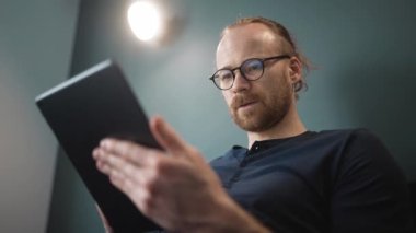 Gözlüklü dalgın sarışın adam evdeki kanepede otururken tablete mesaj atıyor.