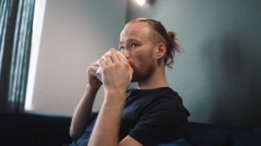 Relaxed blond man wearing blue t-shirt drinking coffee while sitting on the sofa at home