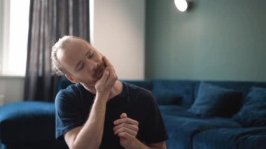 Ciddi sarışın bir adam mavi tişört giyip evinde boynunu yoğuruyor.