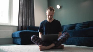 Positive blond man wearing pajama working on laptop while sitting on the floor at home