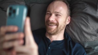 Smiling blond man looking at phone while lying on the bed at home