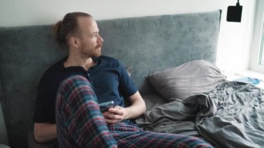 Meditative blond man looking at view and typing on phone while sitting on the bed at home