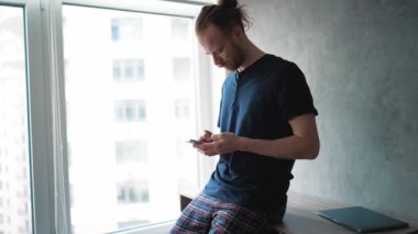 Evde pencerenin yanında otururken telefonla yazan konsantre sarışın adam.