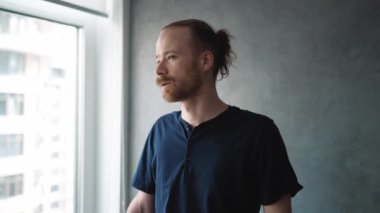 Düşünceli sarışın adam evin penceresinin yanında otururken etrafına bakıyor.