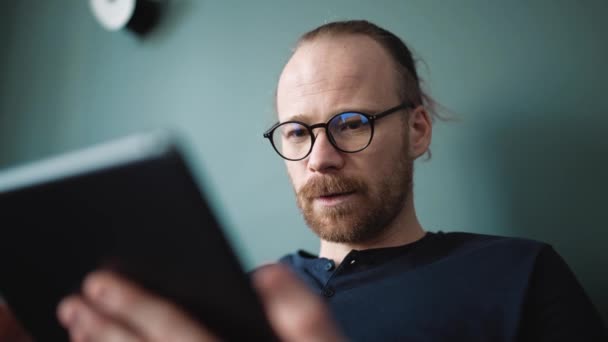 Concentrated Blond Man Wearing Eyeglasses Typing Tablet While Sitting Sofa — Vídeo de Stock