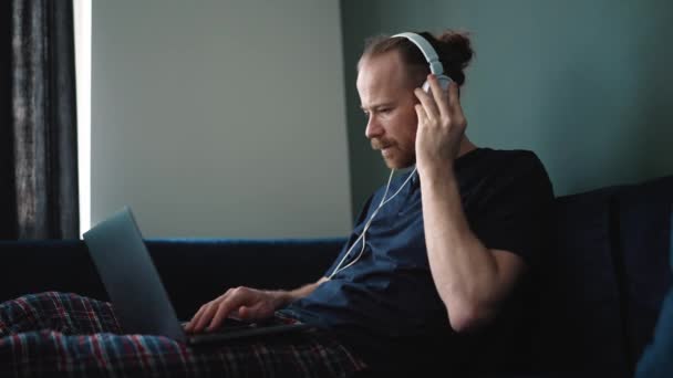 Serious Blond Man Wearing Pajama Working Laptop Headphones While Sitting — Vídeo de stock