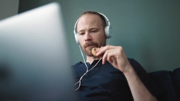 Positive Blond Man Watching Something Laptop Headphones Eating Cookie Home — Stockvideo