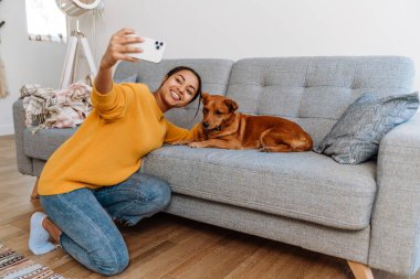 Young hispanic woman taking selfie photo with her dog on cellphone at home
