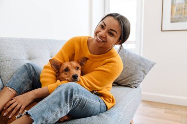 Young hispanic woman smiling while resting on couch with her dog at home