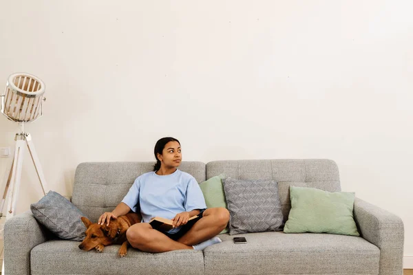 Young Hispanic Woman Petting Her Dog While Reading Book Couch — Fotografia de Stock