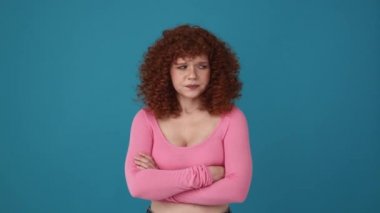 Angry curly haired redhead woman wearing pink shirt showing shh gesture at the camera in blue studio