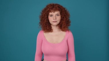 Cheerful curly haired redhead woman wearing pink shirt looking at the camera in blue studio