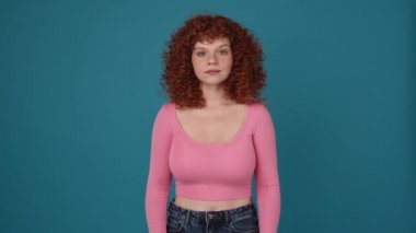 Gülümseyen kıvırcık saçlı, pembe tişörtlü bir kadın mavi stüdyodaki kameraya bakıyor.