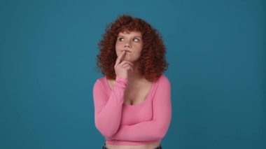 Dalgın kıvırcık saçlı, pembe tişörtlü, mavi stüdyoda düşünen bir kadın.