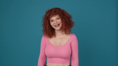 Pretty curly haired redhead woman wearing pink shirt looking at the top in blue studio